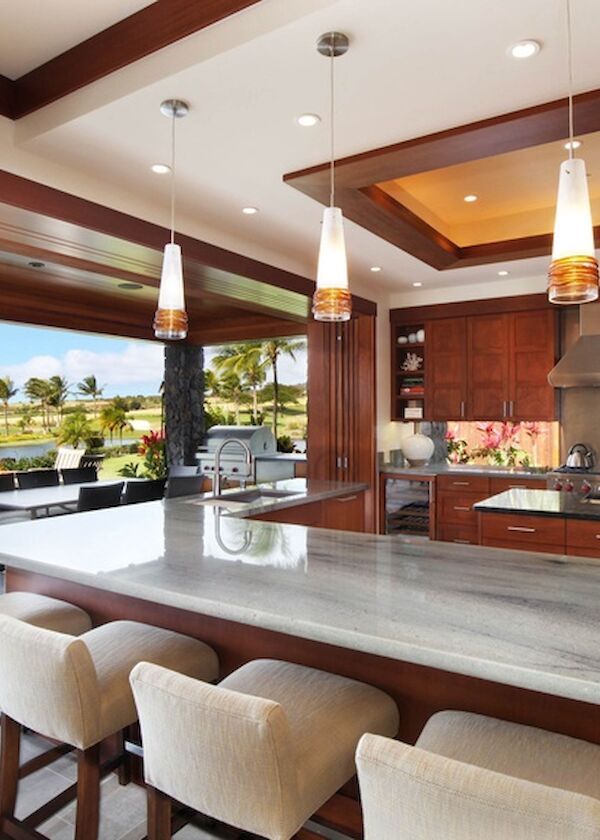 A modern outdoor kitchen with a long marble island, beige bar stools, hanging pendant lights, stainless steel grill, and a covered patio near a lush green landscape.
