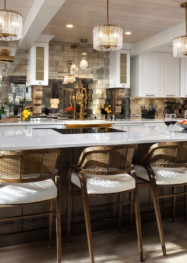 A bright, modern kitchen with white cabinetry, a long marble island, brass bar stools, pendant lights, and stainless steel appliances.