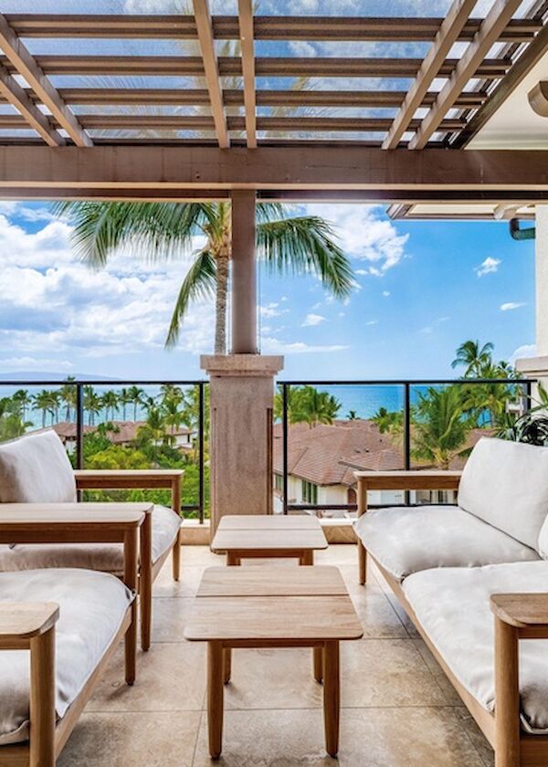 A stylish outdoor patio with white cushioned sofas, wooden chairs, a central coffee table, palm trees, and a sea-view backdrop.