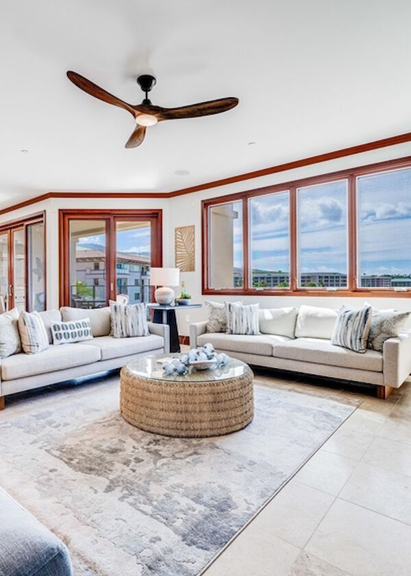 Bright, airy living room with large windows, modern white sofas, a circular coffee table, and a TV; coastal views outside, ceiling fans overhead.