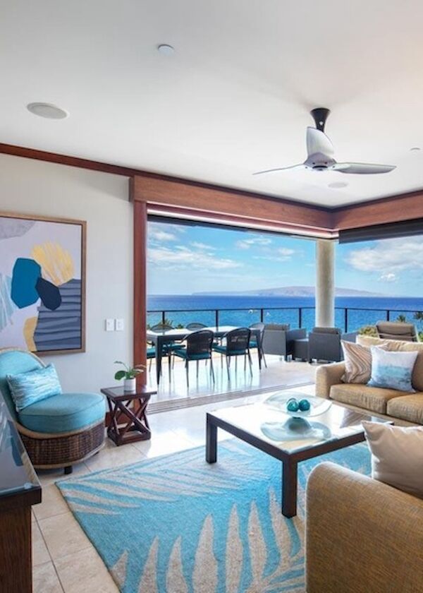 A bright beachfront living room with a bay window view of the ocean, beige sectional sofa, blue accents, and a balcony dining area.