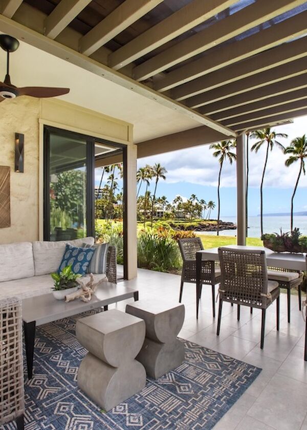 A stylish outdoor living space with a cream sectional, woven furniture, blue patterned rug, ceiling fan, and a dining area overlooking palm trees and the ocean, under a shaded terrace.