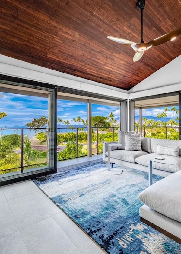 A bright coastal living room with large windows, a light gray sectional, blue rug, wooden ceiling, ceiling fan, and a balcony overlooking greenery.