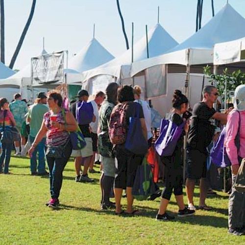 People line up at white tents in an outdoor fair or market on a sunny day, carrying bags and wearing casual clothing, waiting to enter.