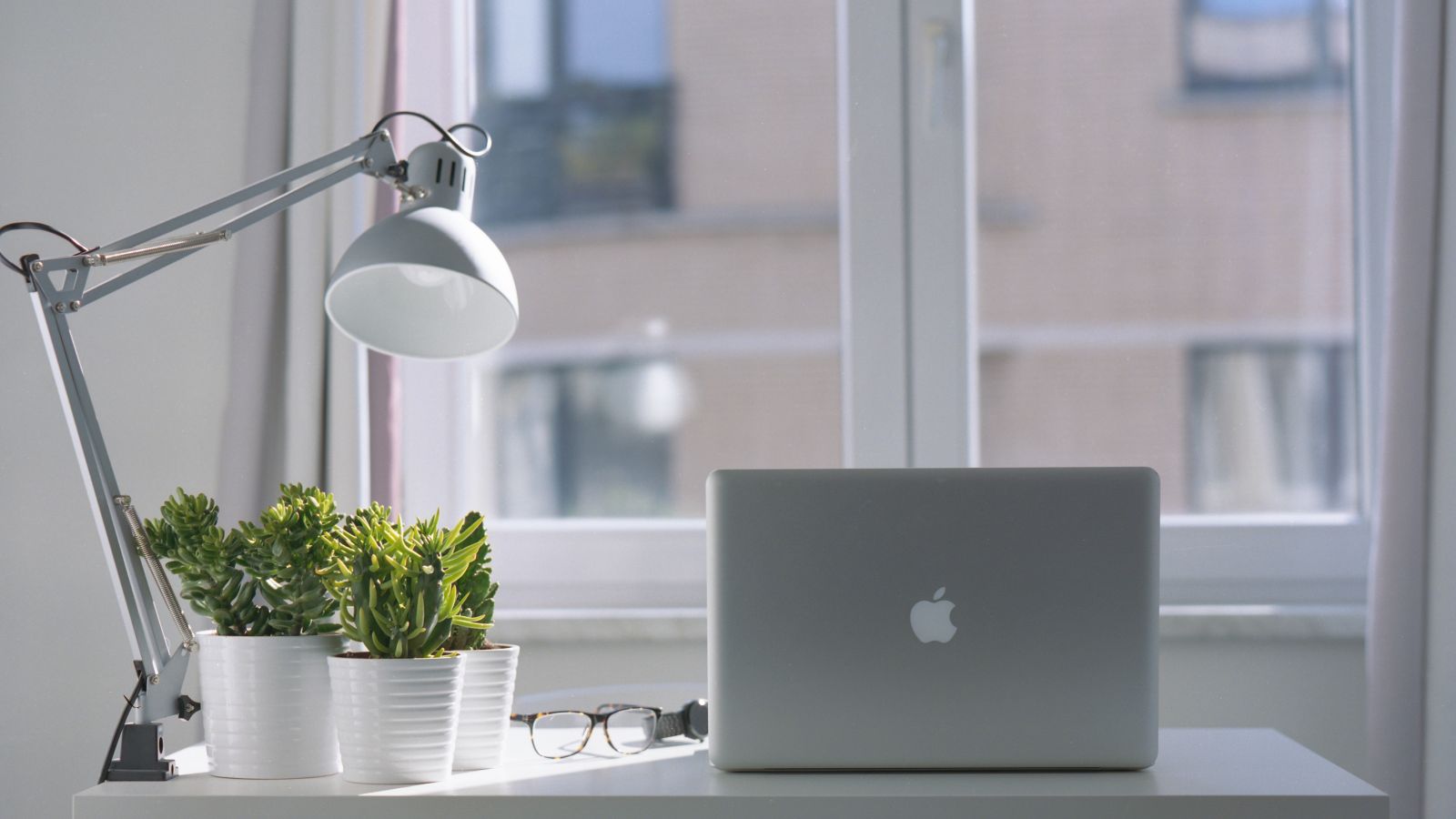 A minimalist desk setup features a laptop, desk lamp, potted plants, and glasses by a window.