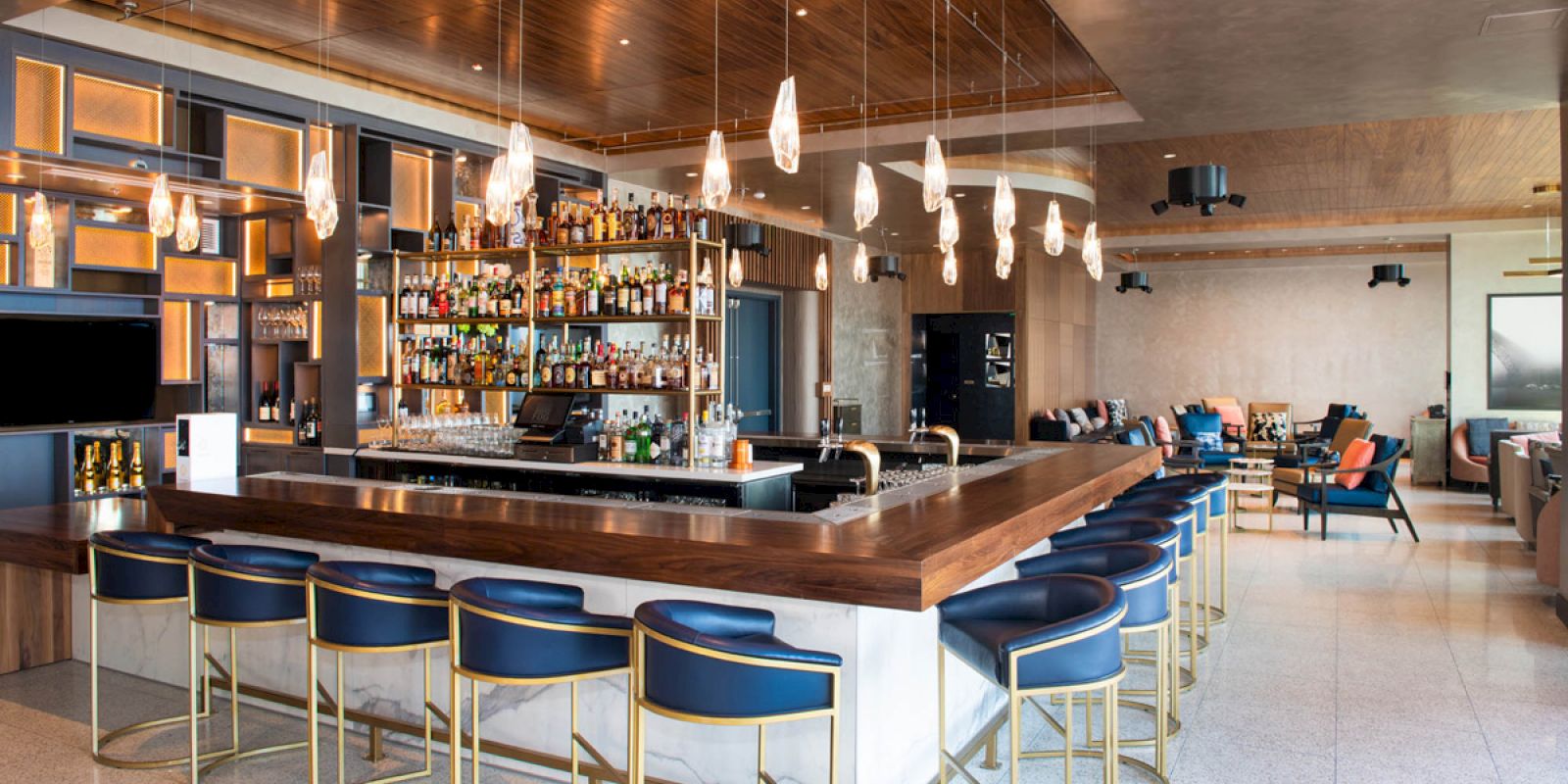 The image shows a modern bar with a sleek, wooden counter, a variety of liquors on shelves, blue barstools, and stylish pendant lights hanging above.