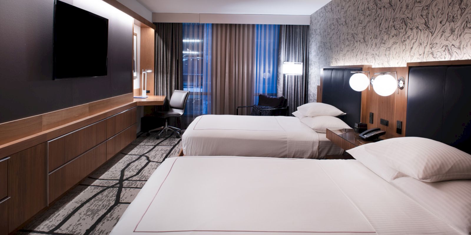 This image shows a modern hotel room with two double beds, a TV, a desk with a chair, and stylish lighting, including lamps by the beds.