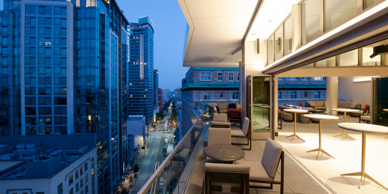 A modern rooftop terrace with seating areas and tables, overlooking a cityscape with tall buildings at dusk, and a brightly lit interior space visible.