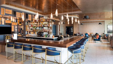 A modern bar area with a wooden countertop, blue bar seats, hanging lights, and a well-stocked liquor shelf, adjacent to a comfortable lounge area.