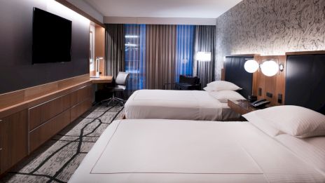 A modern hotel room with two twin beds, a wall-mounted TV, a desk with a chair, lamps, and curtains covering the windows, ending the sentence.