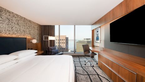 A modern hotel room with a large bed, flat screen TV, desk, chair, and a view of a cityscape through the window, and wall art decorates the room.