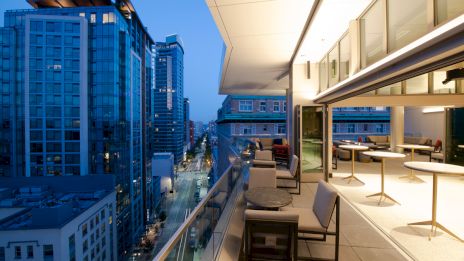 A modern rooftop terrace with seating and tables, overlooking a cityscape with tall buildings at dusk, offering a scenic view of the urban environment.