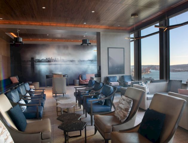 The image shows a lounge area with numerous chairs, tables, large windows, and a mural of a city skyline on the wall.
