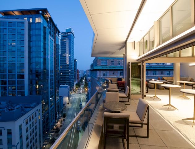 A modern balcony at dusk overlooks a city street with high-rise buildings, featuring a seating area with chairs, tables, and overhead lighting.