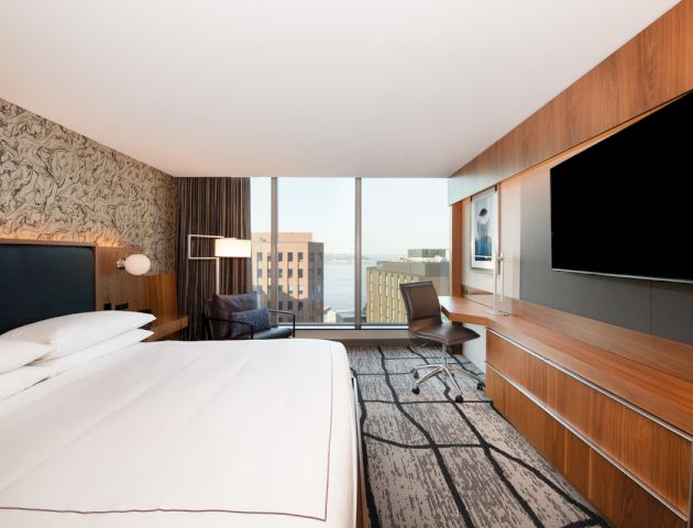 A modern hotel room with a large bed, TV, desk, chair, and a view of buildings and water through floor-to-ceiling windows.