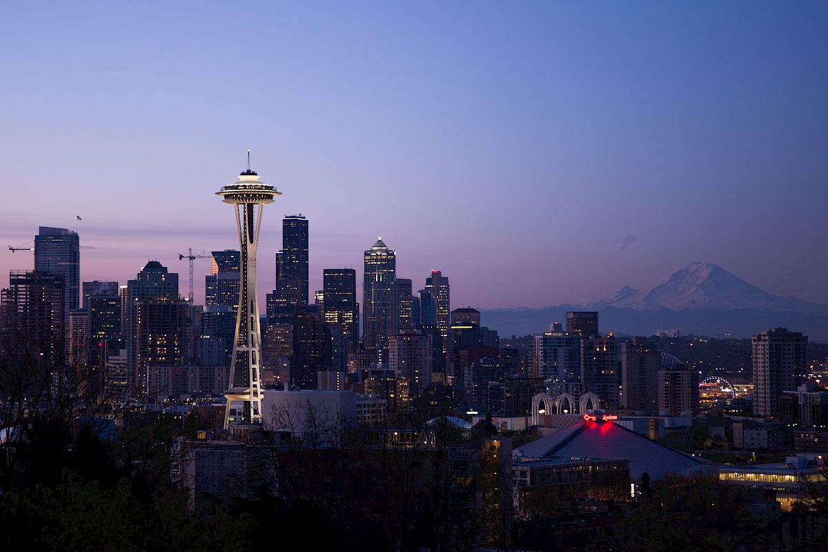 The image shows the Seattle skyline at twilight with the Space Needle prominent, skyscrapers, and Mount Rainier in the background.