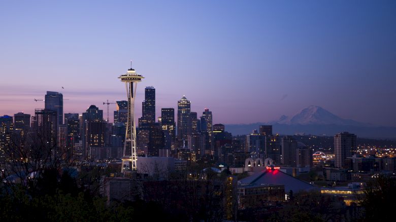 The image shows the Seattle skyline at twilight with the Space Needle prominent, skyscrapers, and Mount Rainier in the background.