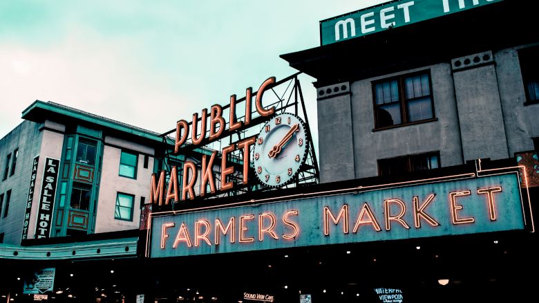 A neon sign reads "Public Market" with a clock and "Farmers Market" below, in a busy market area with surrounding old-style buildings.
