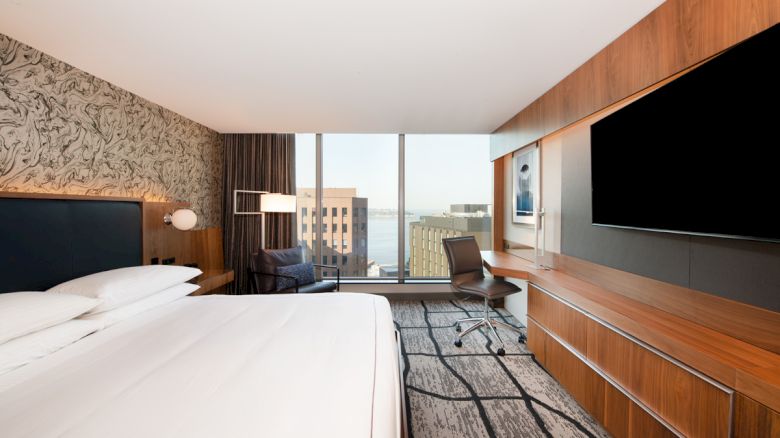 The image shows a modern hotel room with a large bed, a wall-mounted TV, a functional work desk, and a view of the city through large windows.