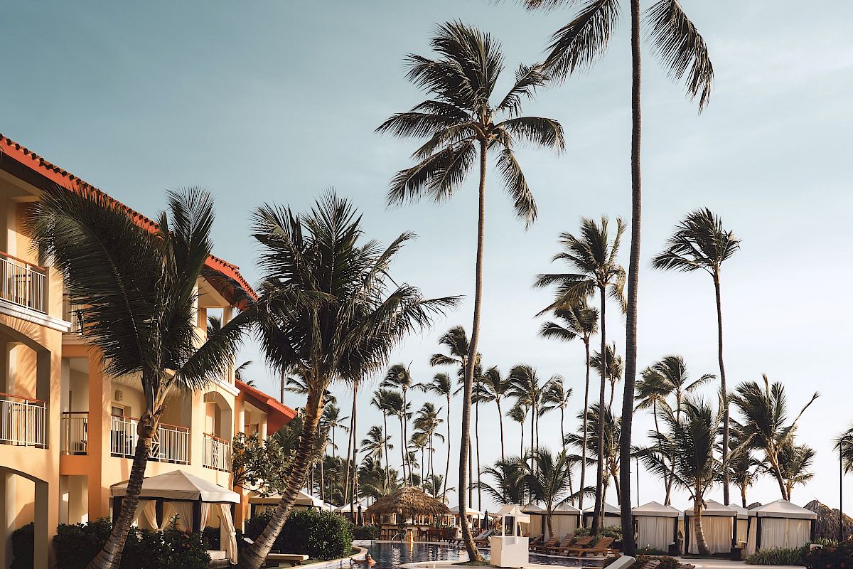 A serene poolside scene with lounge chairs, tall palm trees, and an adjacent building under a clear blue sky, evoking a tropical resort atmosphere.